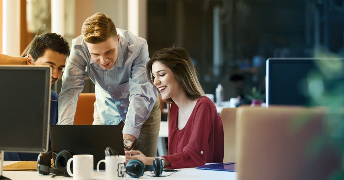 Happy office workers huddle around a laptop computer at a work station.